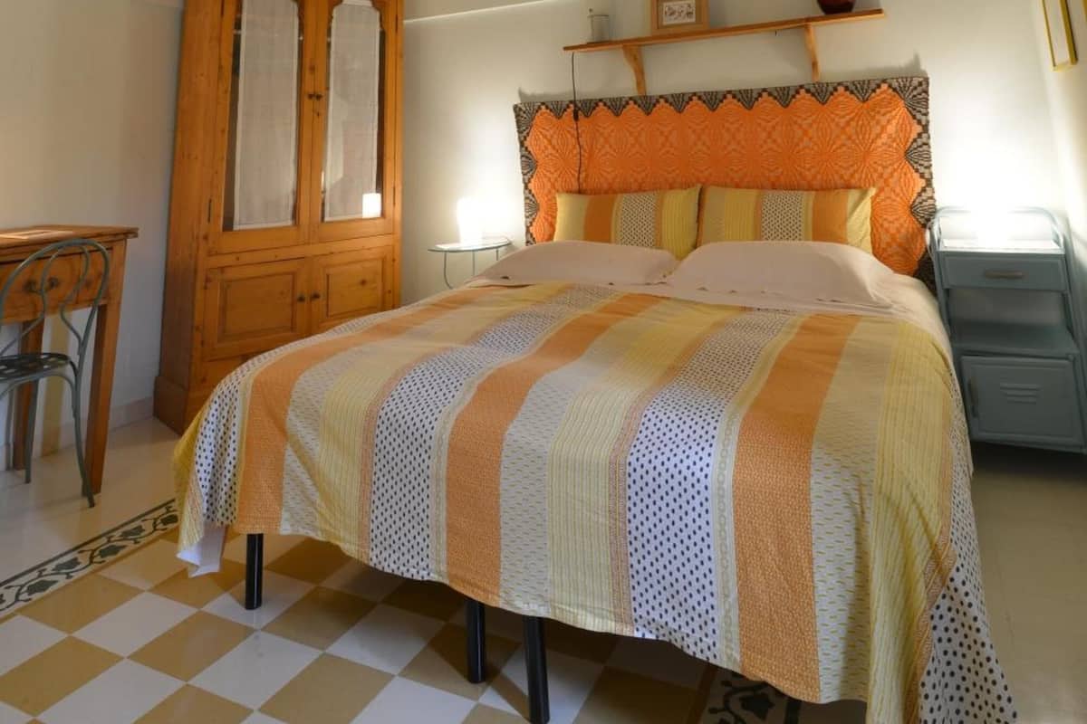 Une chambre avec un sol à carreaux, un lit recouvert d’une couverture à rayures, une armoire en bois et un plafond en briques.