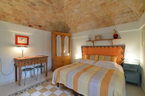 A bedroom with a brick ceiling, a wooden desk with chairs, a bed with striped bedding, and a nightstand with a lamp.