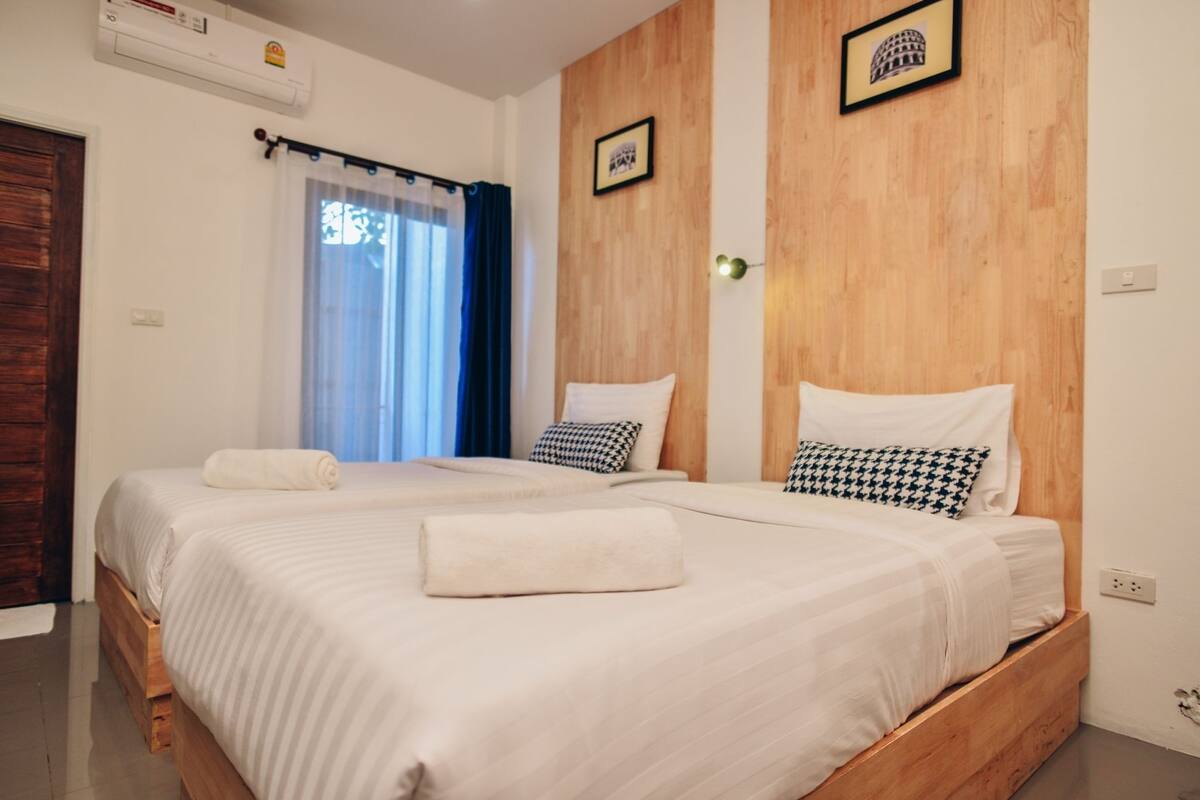 Cama doble con sábanas blancas y dos almohadas, cabecera de madera, ventana con cortinas azules y un cuadro en la pared.
