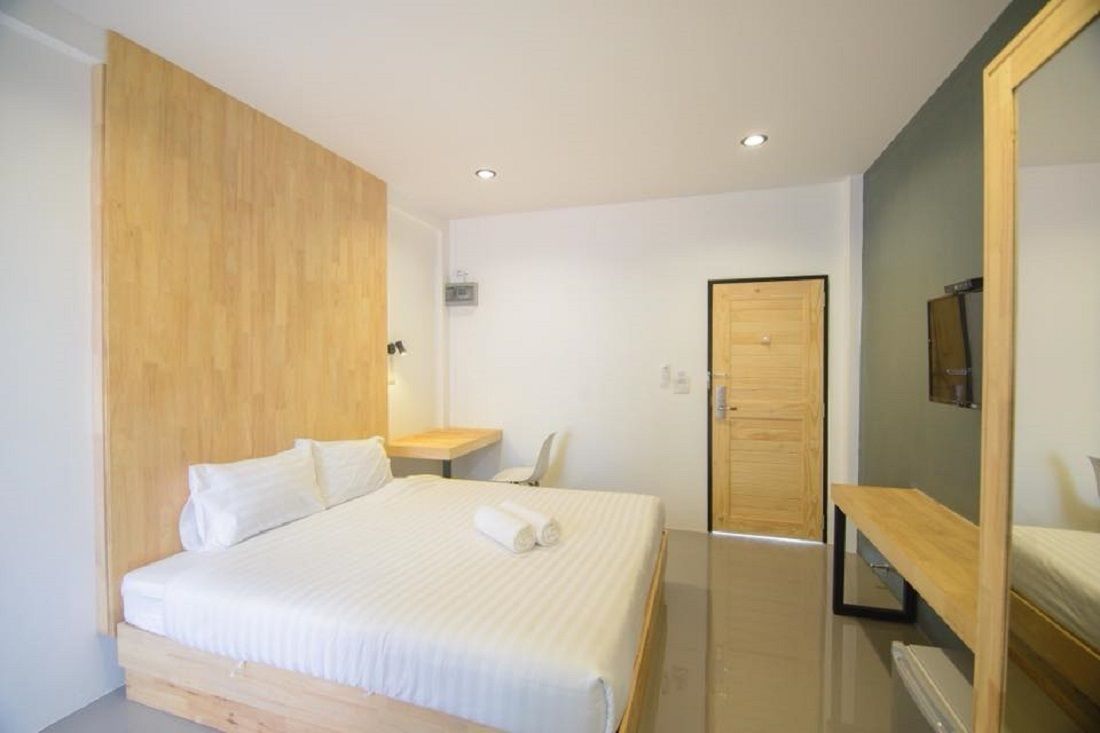 Una habitación de hotel moderna con un cabecero de madera, una cama con ropa blanca, una mesita de noche, una silla y un espejo.