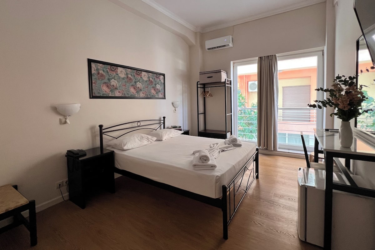 A bedroom with a bed, bedside table, wall-mounted air conditioning unit, and a view of the outside balcony.