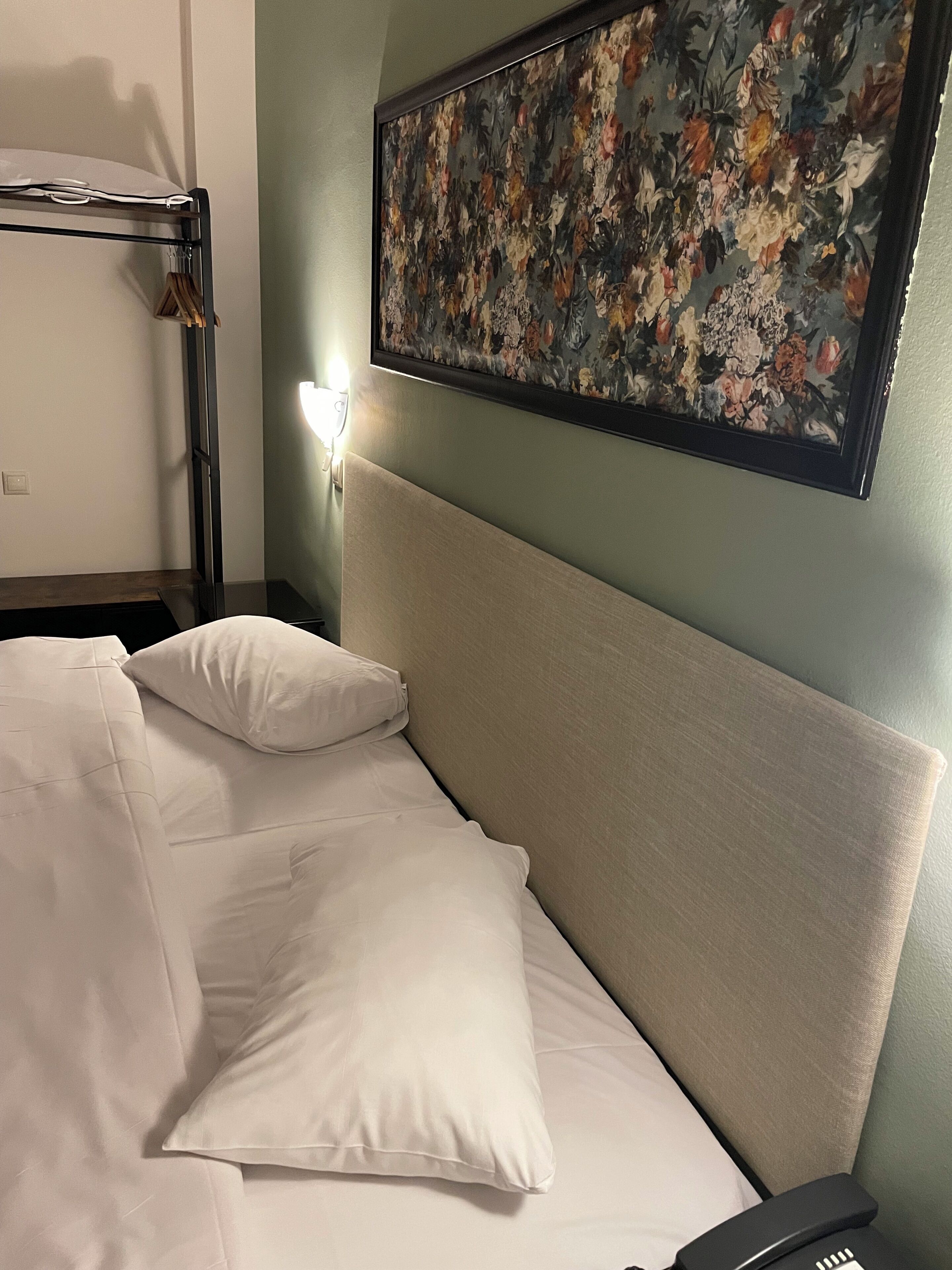 A hotel room with a bed, pillows, a nightstand, a lamp, and a floral painting on the wall.