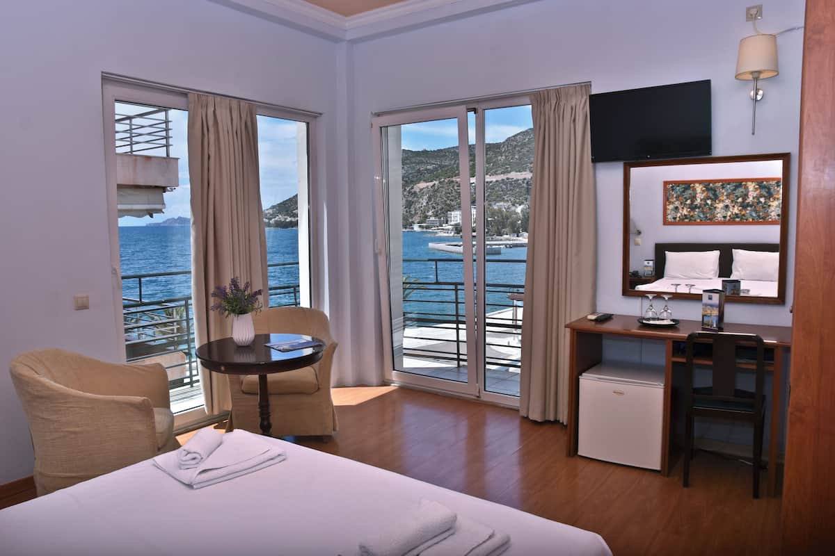 A hotel room with a bed, a desk, a chair, a small fridge, a TV, and a balcony with a view of the sea.
