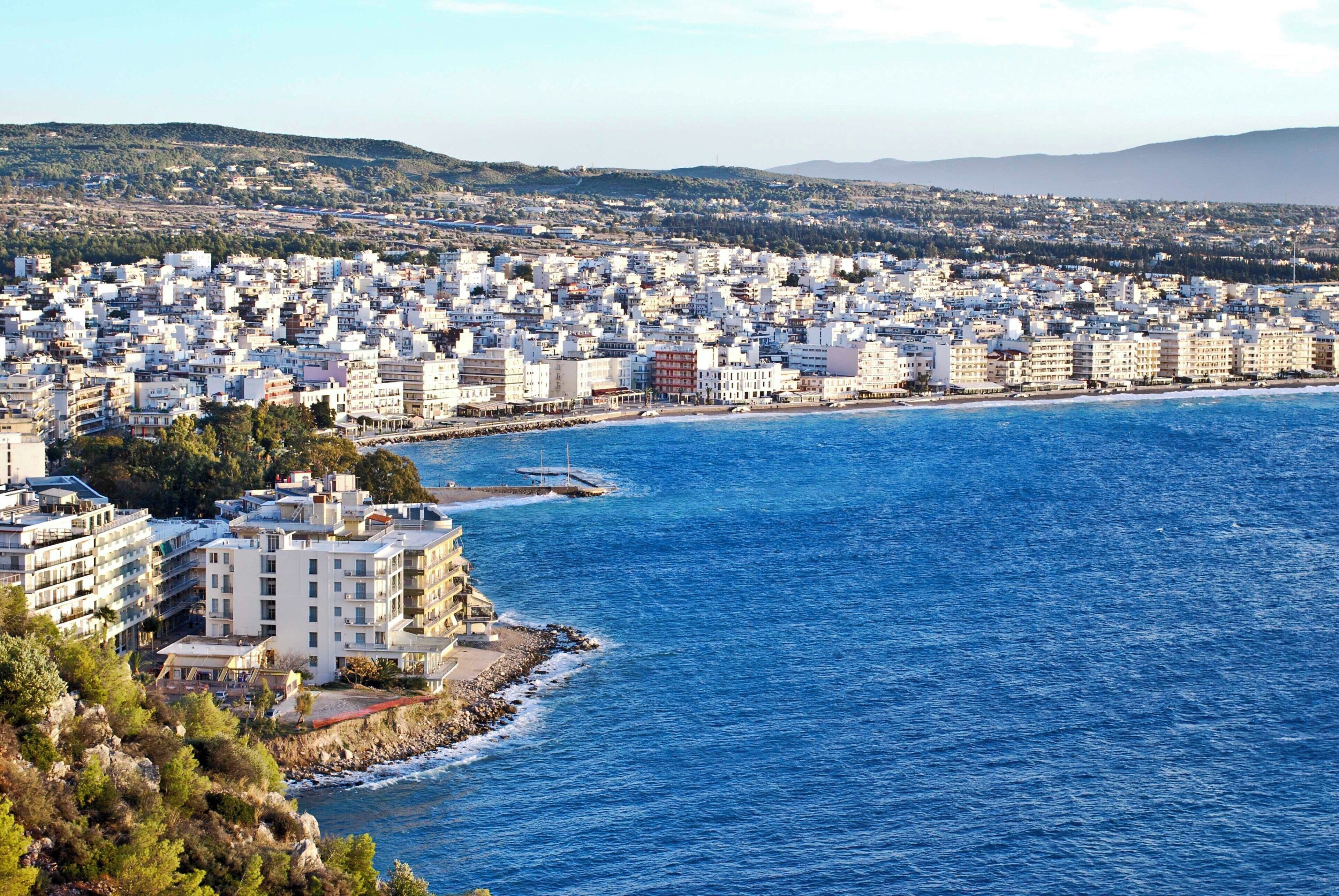 A coastal cityscape with densely packed buildings, a clear blue sea, and a hilly background.