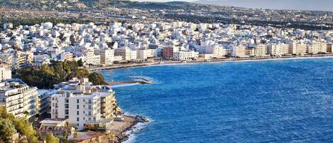 Un paisaje urbano costero con edificios muy juntos, un mar azul cristalino y un fondo montañoso.