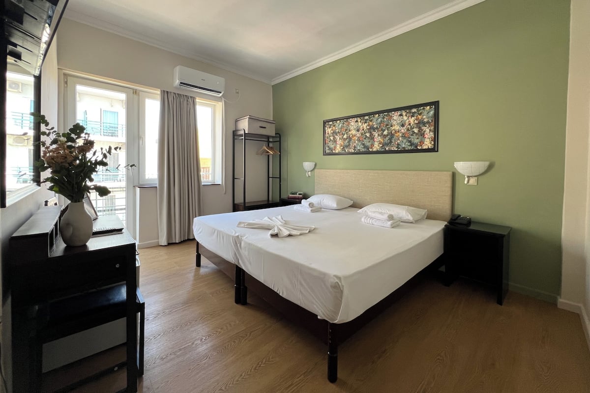 A hotel room with a bed, a desk, a chair, a vase with flowers, a painting on the wall, and an air conditioning unit.