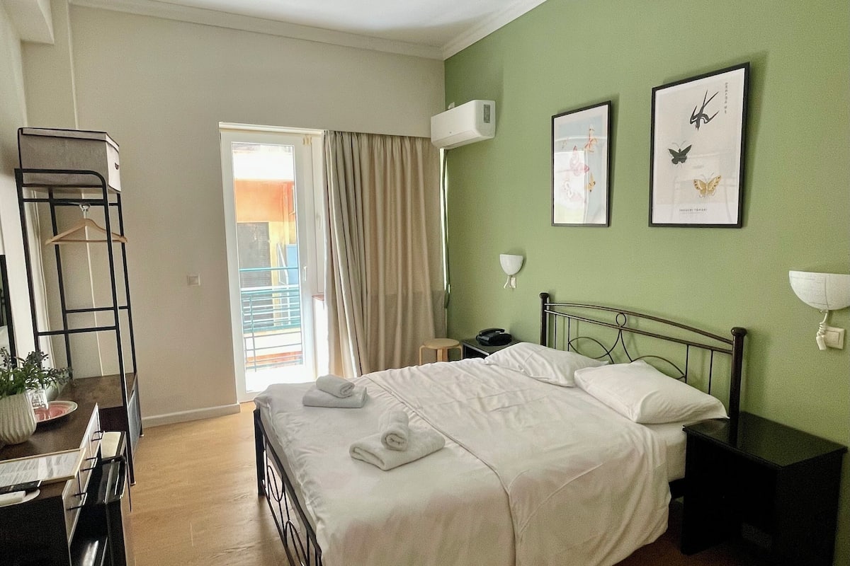 A hotel room with a bed, bedside table, wall-mounted air conditioner, and a desk with a chair.