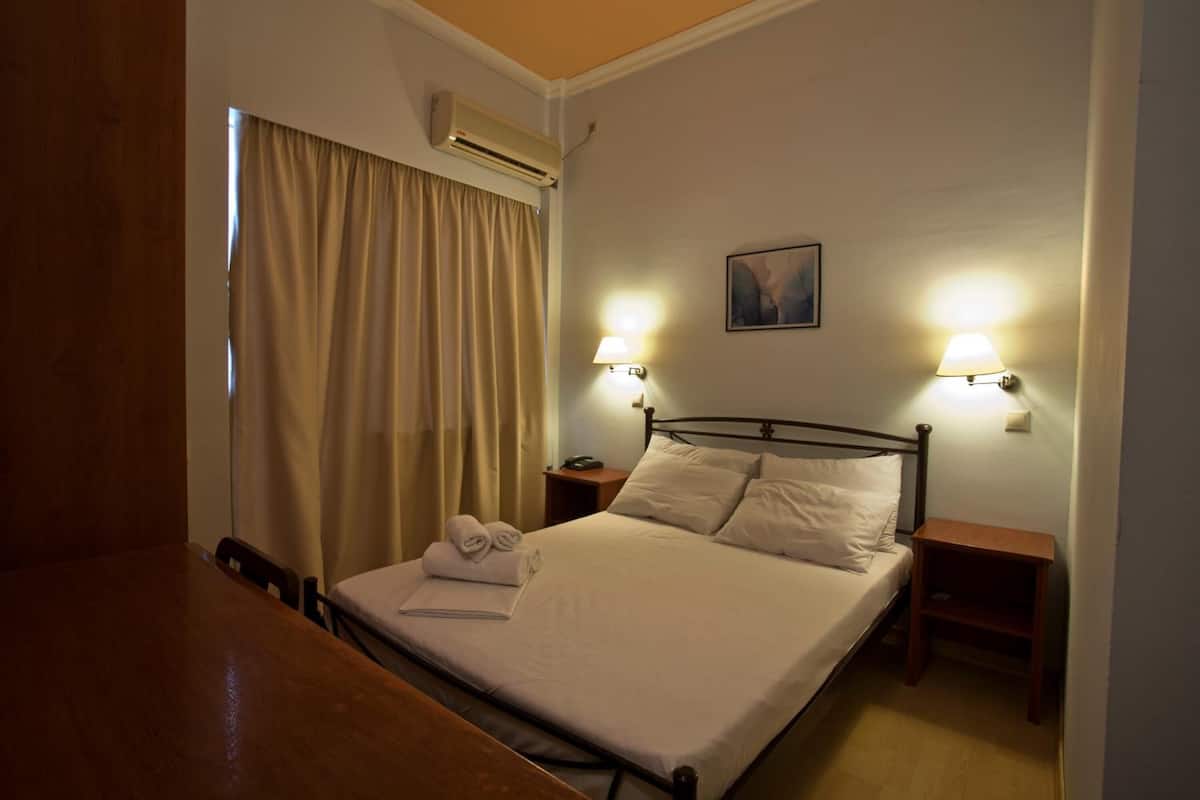 A neatly made bed with white linens, two bedside tables with lamps, a wall-mounted air conditioner, and a window with curtains.