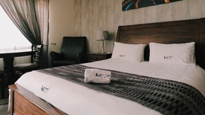 Quarto de hotel com cama grande, cabeceira, escrivaninha com cadeira, abajur e janela com cortinas.
