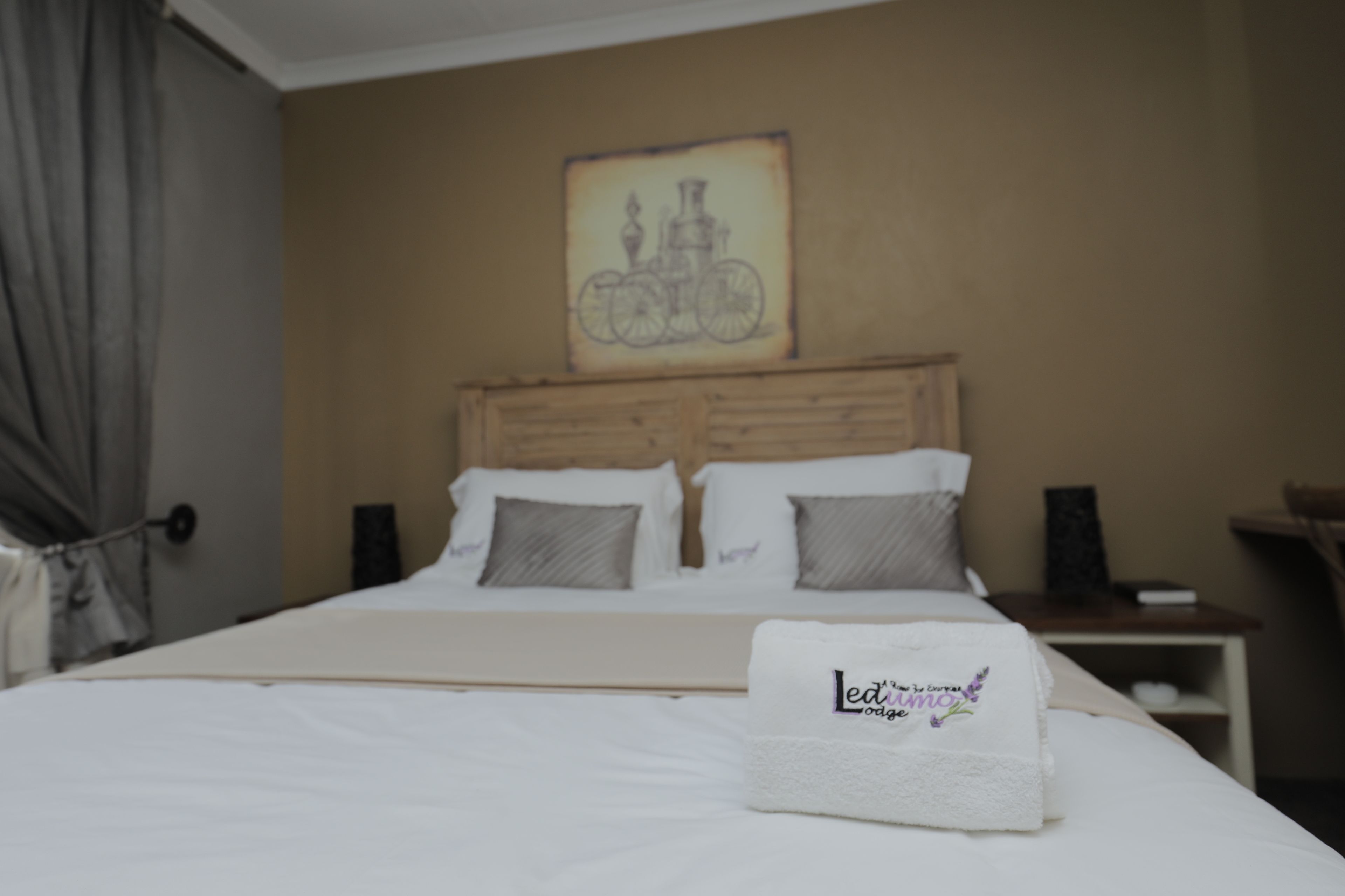 A neatly made bed with a white blanket and a towel with text, a wooden headboard, a wall-mounted artwork of a bicycle, and a bedside table with a book and a lamp.