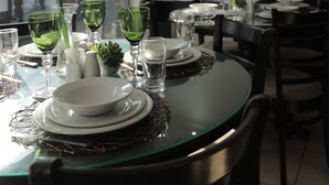 Uma sala de jantar com uma mesa de vidro posta para refeição, incluindo pratos, tigelas e copos. Há uma pequena planta na mesa e o ambiente possui um design moderno, com uma televisão de tela plana fixada na parede.