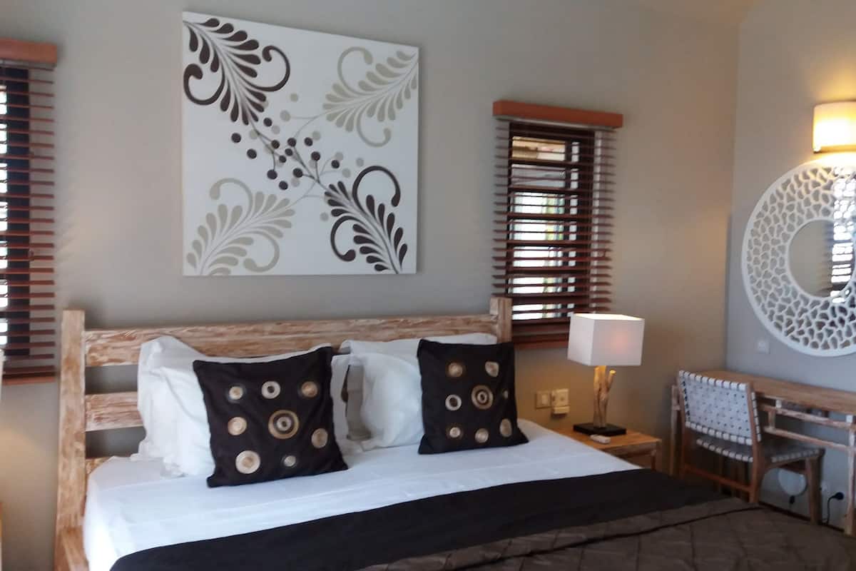 A hotel room with a large bed, two pillows, a bedside table, a lamp, a chair, a decorative wall piece, and a round mirror.