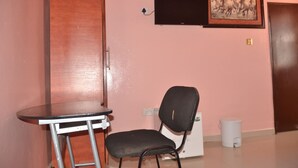 A room with a wooden wardrobe, a small table with a chair, a television mounted on the wall, and a framed picture above the TV.