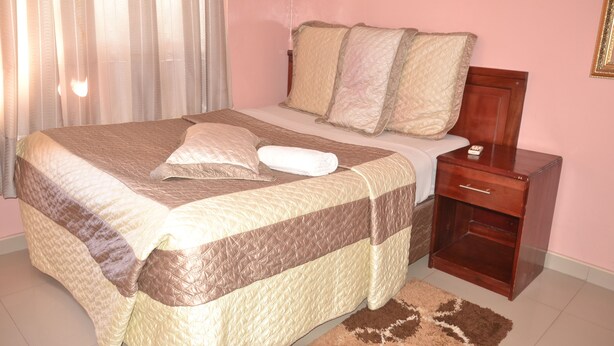 A neatly made bed with a brown and beige quilt, a wooden nightstand with a drawer, and a brown rug on a tiled floor.