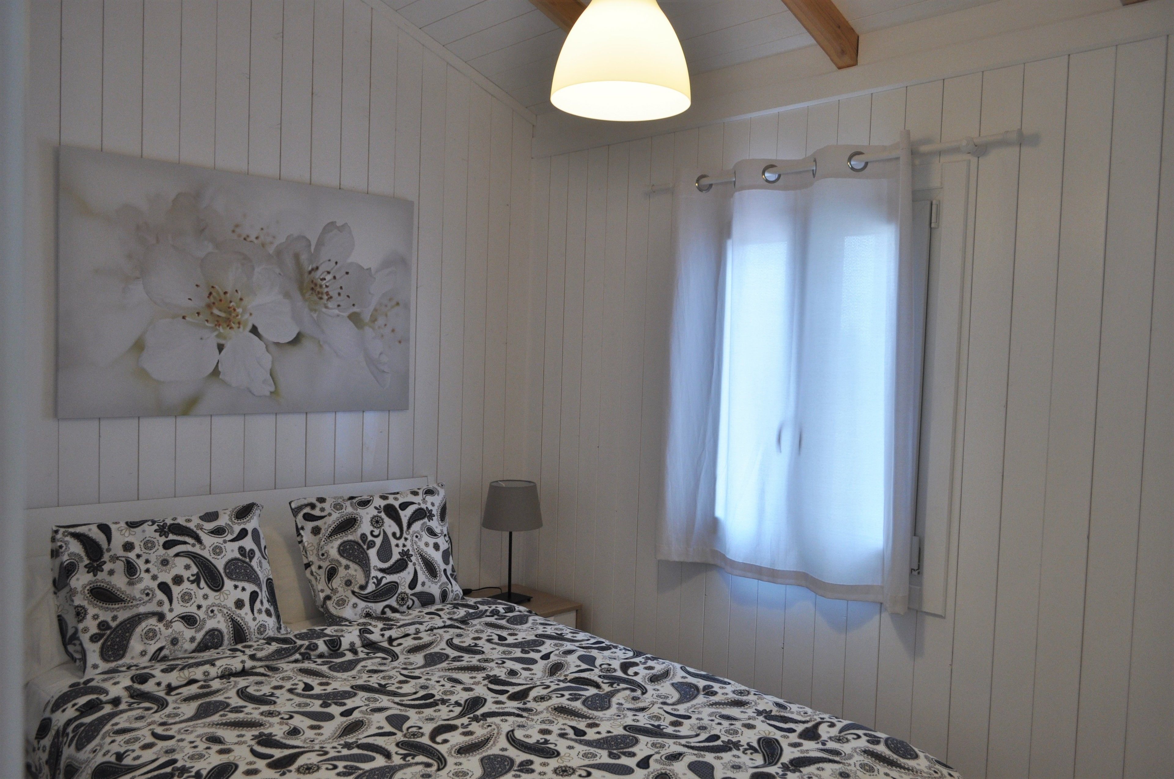 Ein Schlafzimmer mit einem Bett, einem Nachttisch, einer Lampe, einem Fenster mit Vorhängen und einem Wandbild einer Blume.