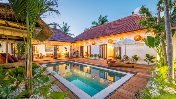 Un espace piscine doté d’une terrasse en bois, entouré de plantes tropicales et bordé d’un bâtiment dont le toit est recouvert de tuiles.