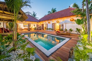 A pool area with a wooden deck, surrounded by tropical plants and a building with a tiled roof. - Villa Makasih by Villa Finder (Seminyak)