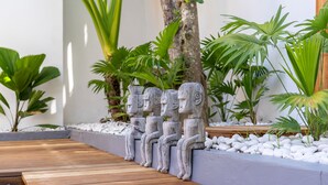 Une terrasse en bois ornée de statues en pierre, entourée de plantes tropicales et bordée d’un mur blanc.