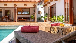 Un espace aménagé au bord de la piscine, avec un fauteuil de relaxation en osier, une serviette rouge et une table en bois sur laquelle se trouve un panier de fruits.