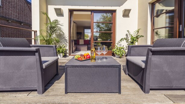 Um pátio externo com móveis de vime, uma mesa com frutas e uma garrafa, e plantas em vasos.