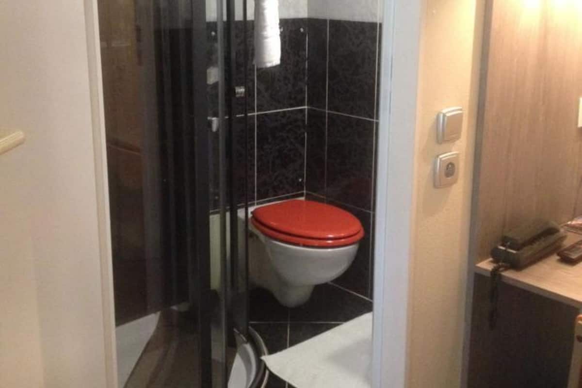 Un baño con cabina de ducha de vidrio, un inodoro con asiento rojo y un pequeño mostrador con un teléfono y documentos.