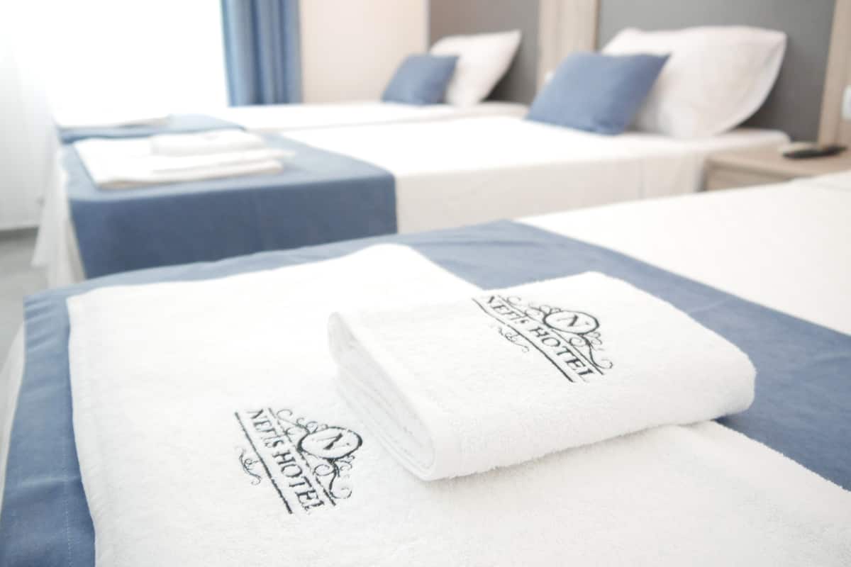 A hotel room with two beds, each with a blue headboard and white bedding. There are towels with the hotel's logo on them.