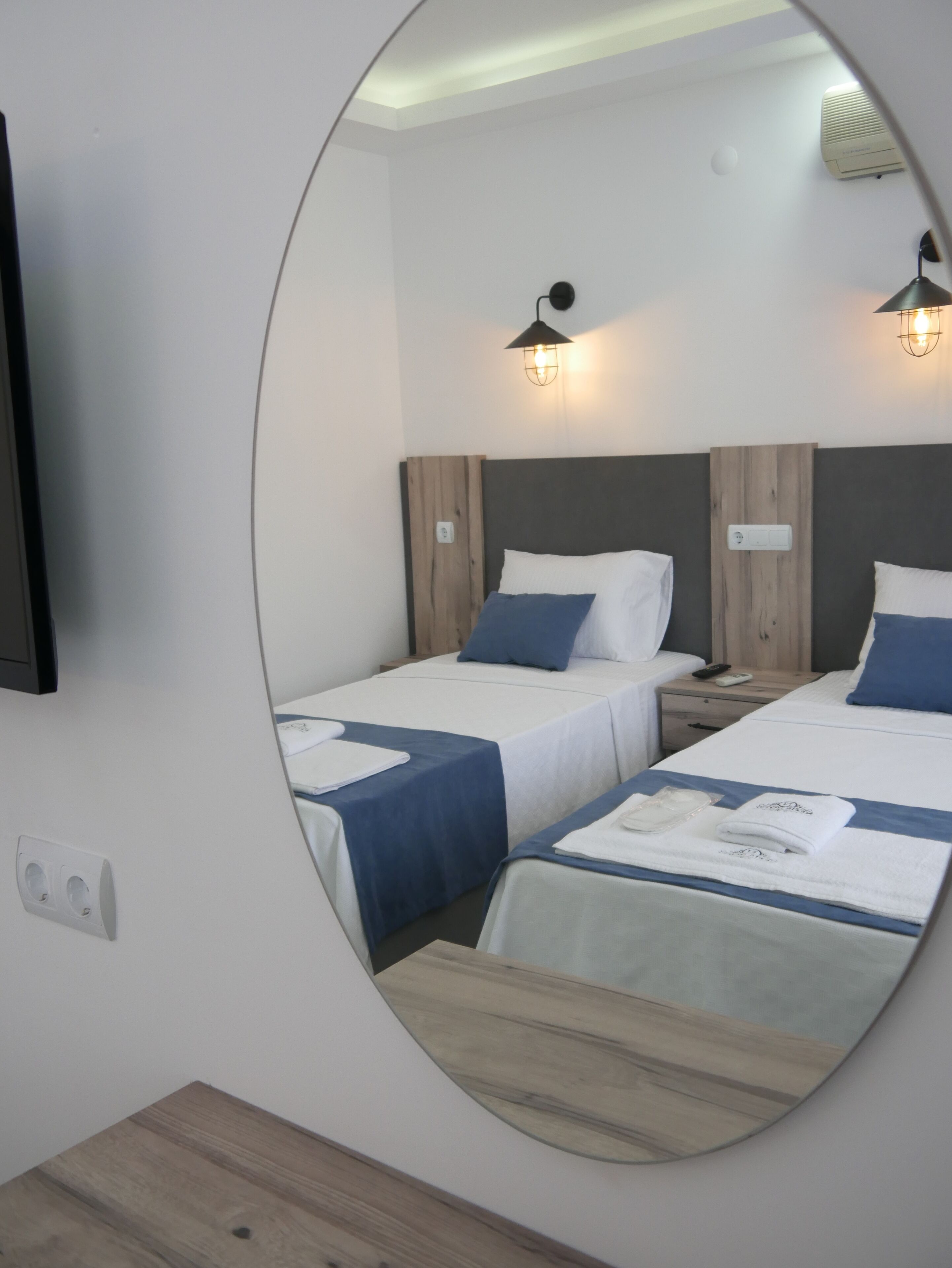A hotel room with two beds, each with a blue bedspread and white pillows. There are wall-mounted lights above each bed, and a flat-screen TV mounted on the wall.