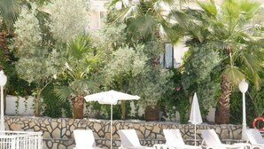 A poolside area with white lounge chairs and umbrellas, surrounded by lush greenery and a stone wall.