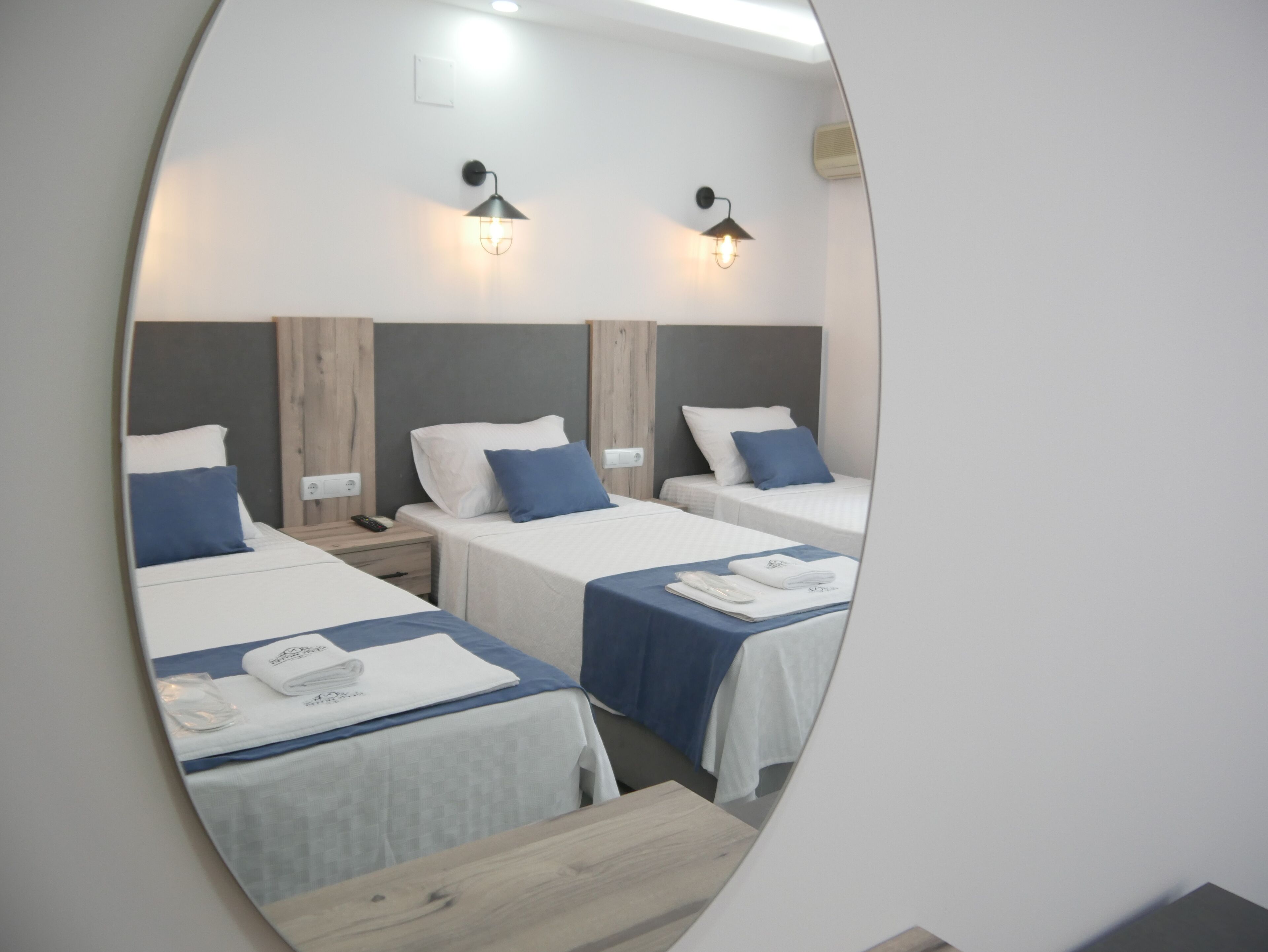 A mirrored view of a hotel room with three beds, each with a blue and white bedspread, a wooden headboard, and a nightstand with a phone and a remote control.