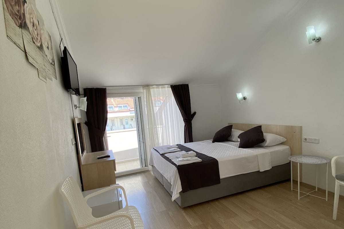 A bedroom with a bed, a small table, a chair, and a desk. There is a window with curtains, a wall-mounted TV, and a light fixture.