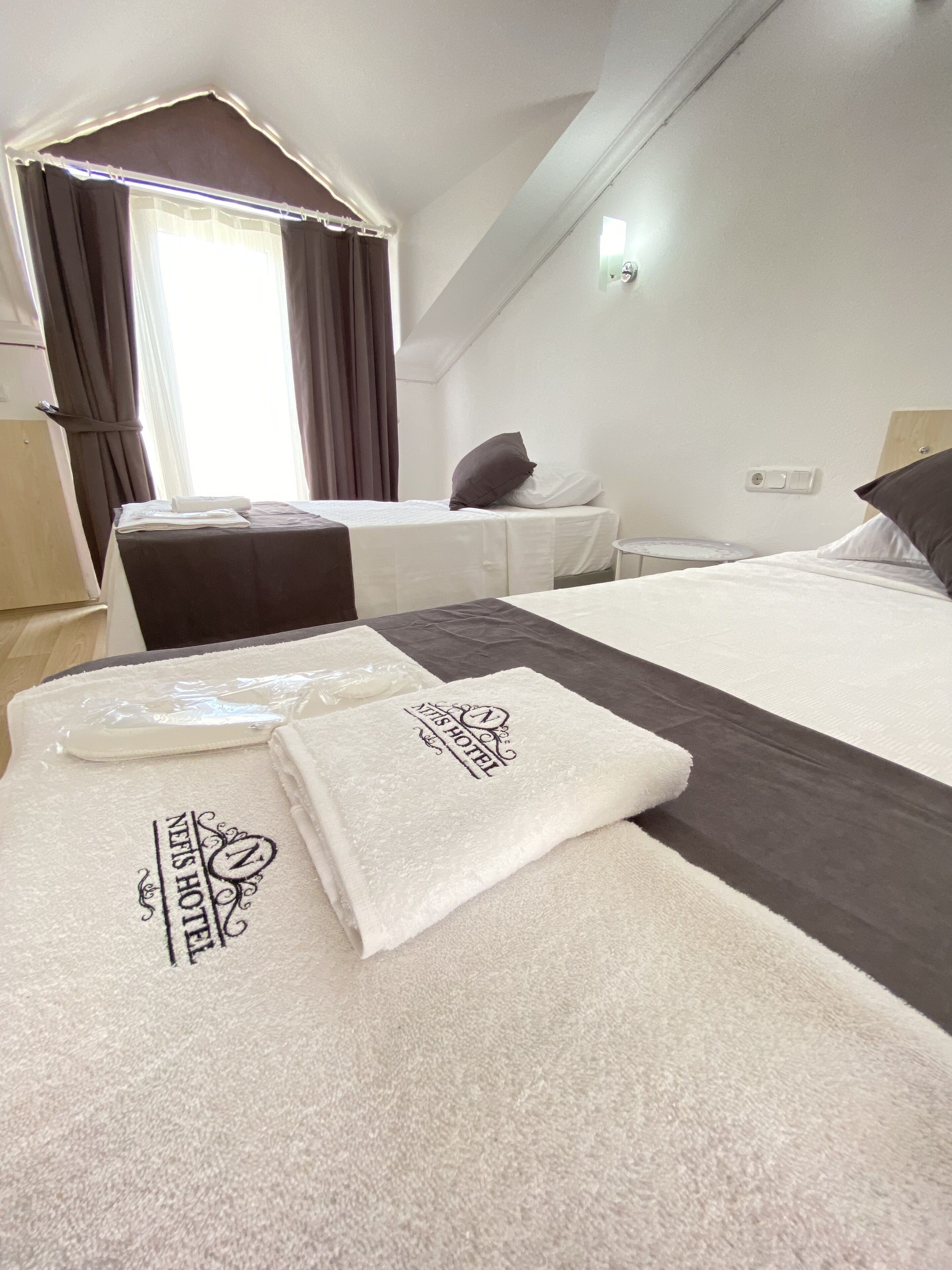 Habitación de hotel con dos camas, una mesita, una ventana con cortinas y una toalla con el nombre del hotel.