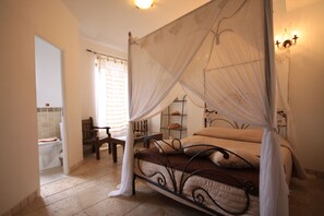 A bedroom with a four-poster bed, a bedside table, a chair, and a shelf with toiletries. - Aux Bords Du Temps (Zonza)