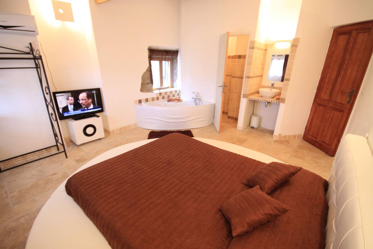 A bedroom with a large bed, a television, a bathroom with a bathtub, and a wooden door.