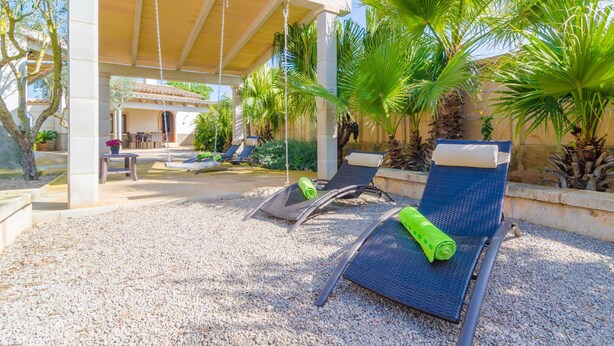 Un espacio de estar al aire libre cubierto, con sillones reclinables y una mesa, rodeado de palmeras y una pérgola.