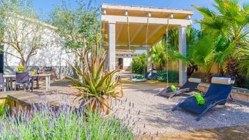 Un espacio de estar al aire libre cubierto con mesas y sillas, rodeado de plantas y una pequeña piscina.