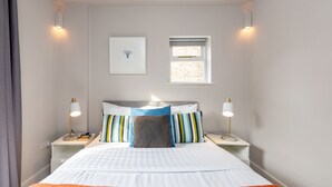 A neatly arranged hotel room with a bed, two bedside tables, a nightstand, a framed picture, and a window with blinds. - Vauxhall Park Views - 2 Bed Flat by BaseToGo (London)