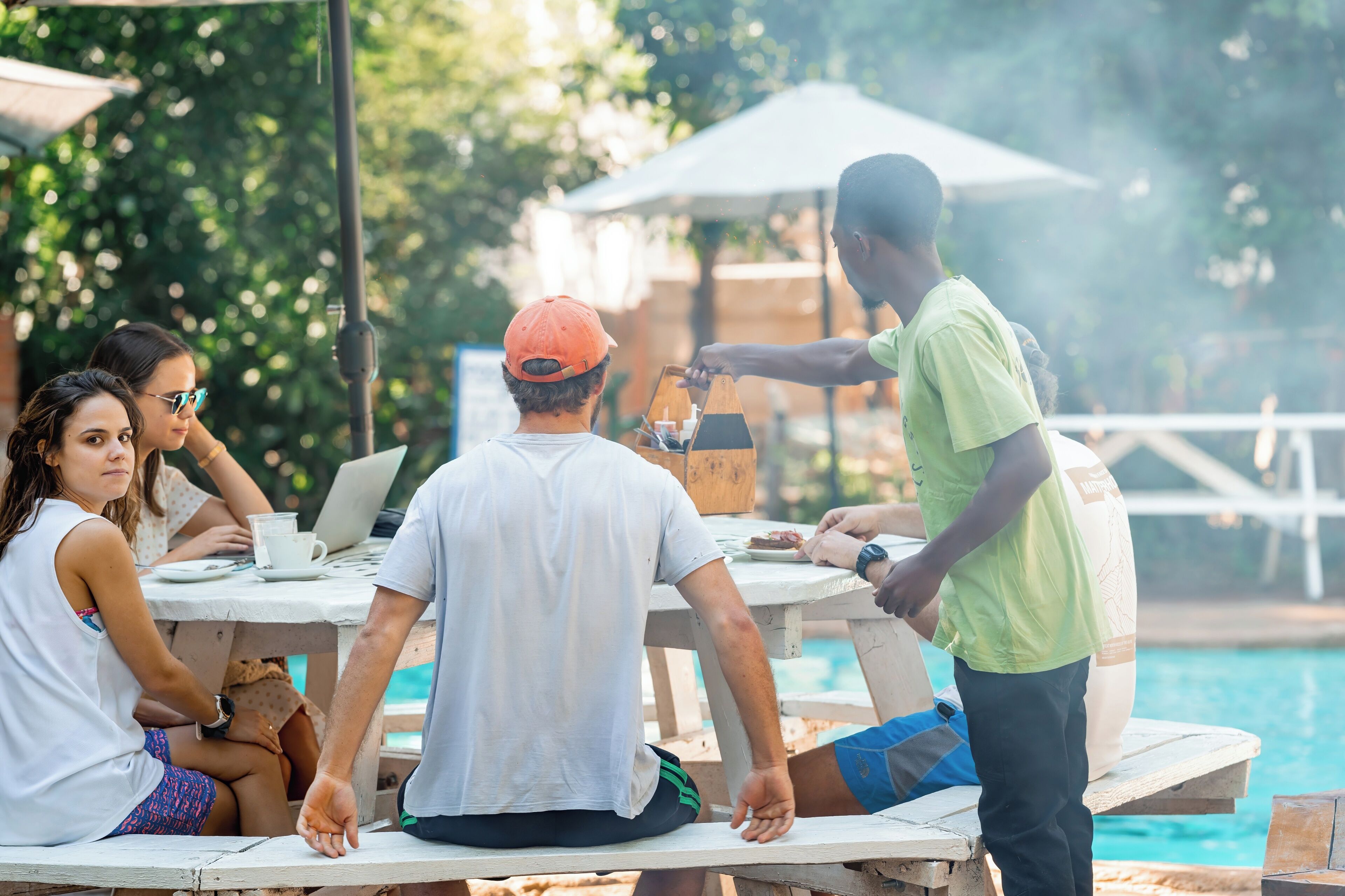 Een groep mensen rond een tafel bij een zwembad, met een barbecue en een laptop.