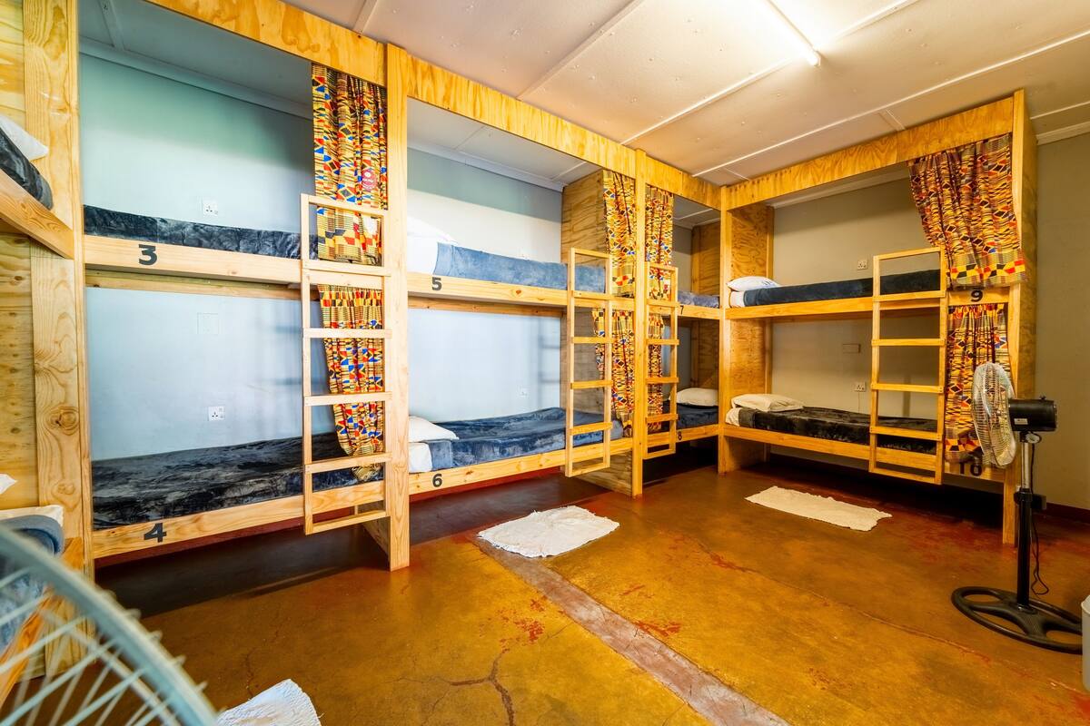 A dormitory room with bunk beds numbered 3 to 9, wooden walls, and a fan in the corner.