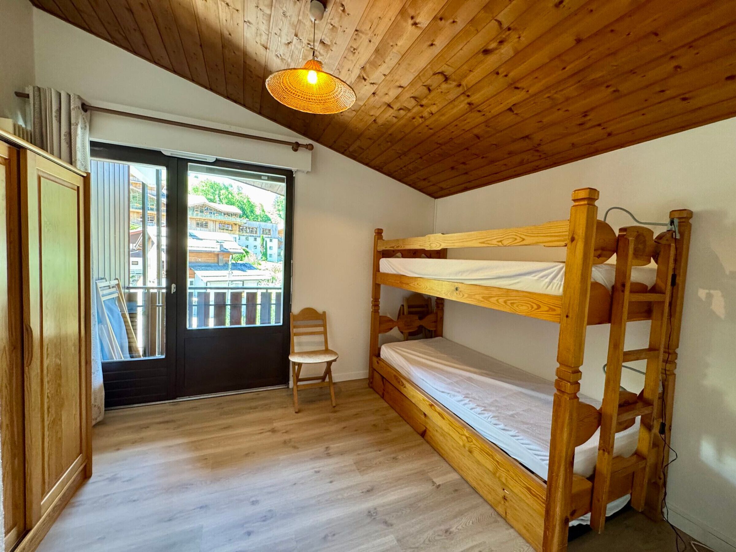 A room with a bunk bed, a chair, a wardrobe, and a window with a view of buildings.