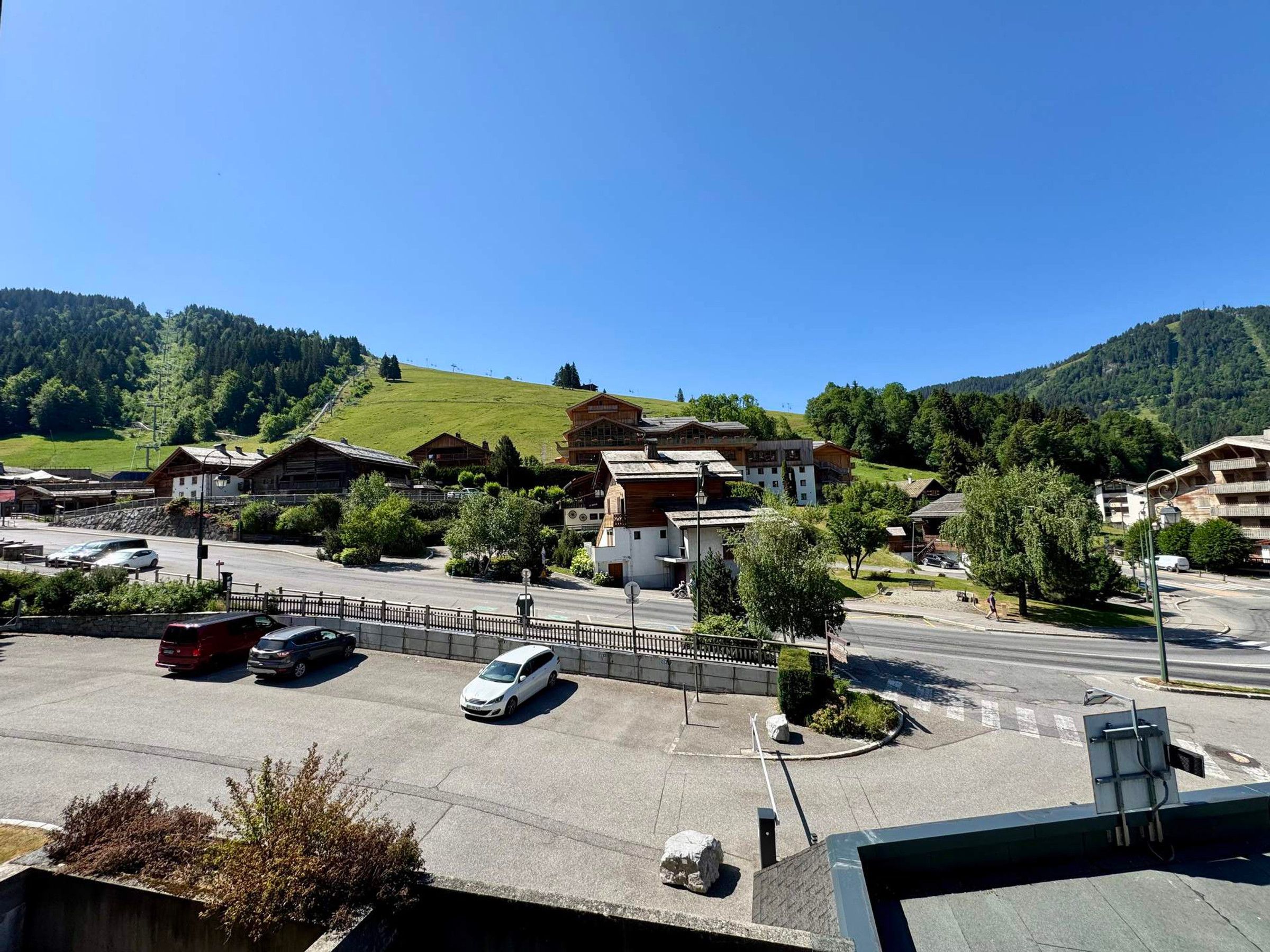 A parking area with cars, a pedestrian crossing, and buildings with a hilly backdrop.