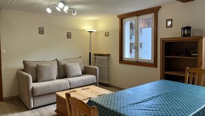 Una habitación acogedora con sofá, mesa de madera, cama con colcha azul, ventana con vistas y un radiador.