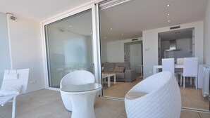 Un espace de vie moderne doté d’une porte-fenêtre coulissante donnant sur un balcon, de meubles en osier blancs et d’une cuisine visible à travers la porte.