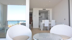 Un salon moderne avec une table en verre et des fauteuils en osier blanc, un coin repas avec des chaises blanches, et une vue sur la mer à travers de grandes fenêtres.
