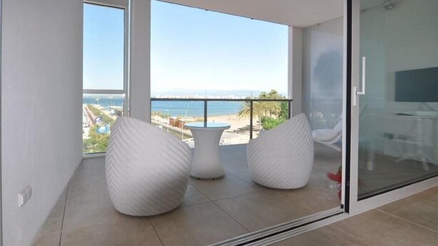 Un balcon moderne avec des sièges rembourrés blancs et une porte en verre, offrant une vue sur une plage et le paysage urbain.