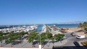 Un port de plaisance où sont amarrés de nombreux bateaux, un parking rempli de voitures et une plage bordée de palmiers.