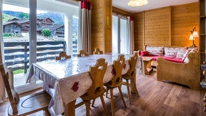 A wooden interior with a dining table, chairs, a sofa, and a view of a village through large windows.