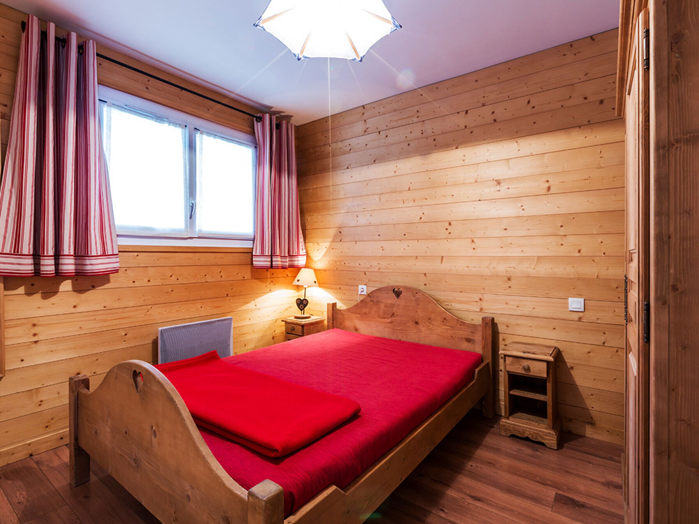 A wooden room with a bed, a window, curtains, a lamp, and a small wooden cabinet.
