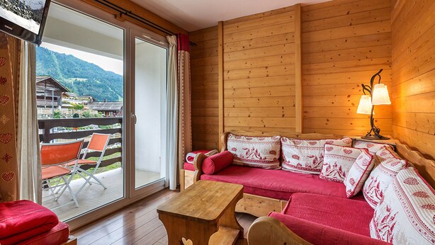 A cozy room with a wooden interior, a sofa with patterned cushions, a wooden coffee table, and a view of a mountain landscape through a sliding glass door.