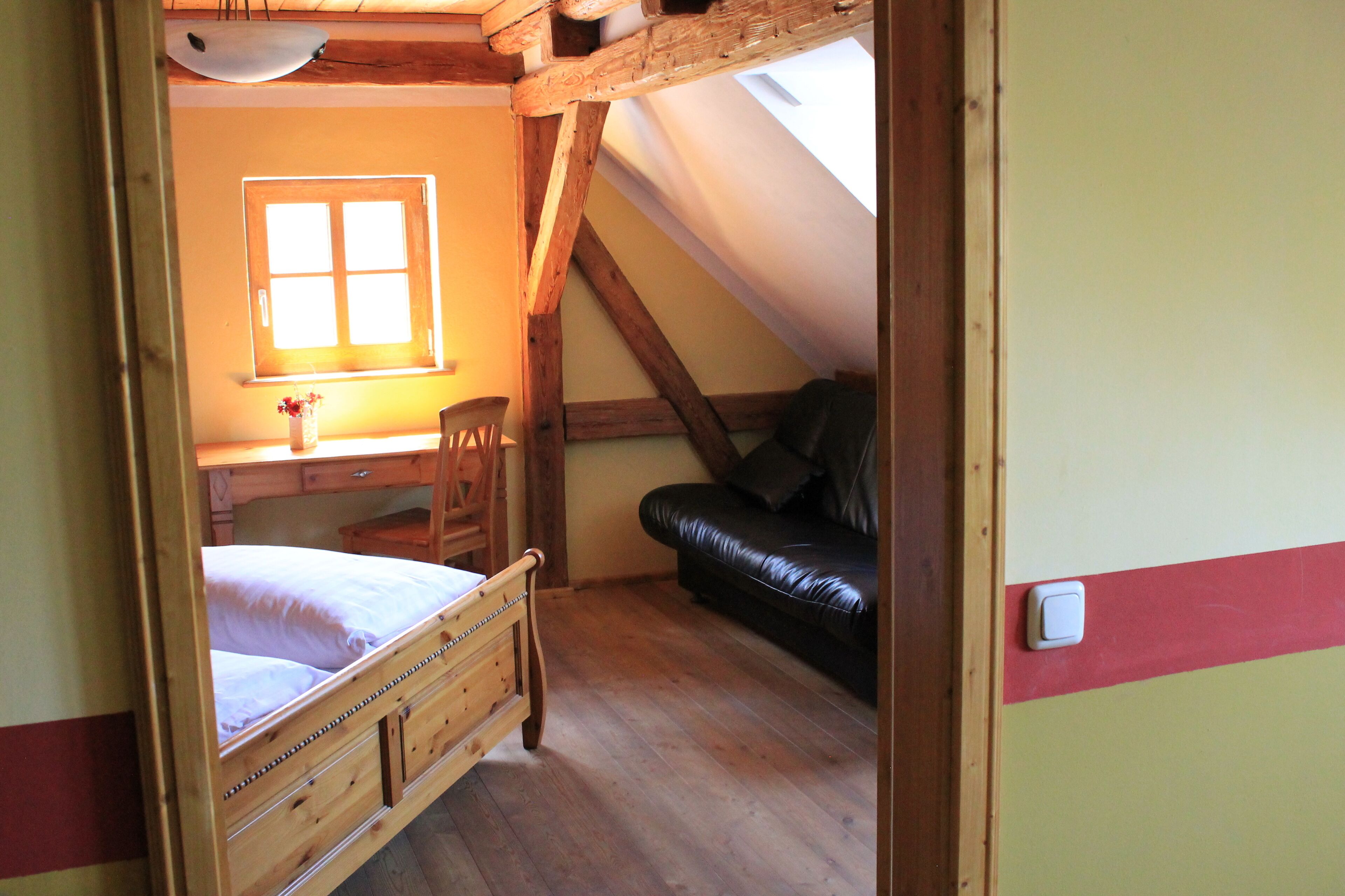 Un dormitorio con una cama de madera, un escritorio con una silla, un sofá de cuero negro y una ventana con luz natural.
