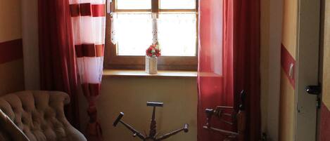 Una habitación con piso de madera, un sillón capitonado, un rueca y una ventana con cortinas rojas.